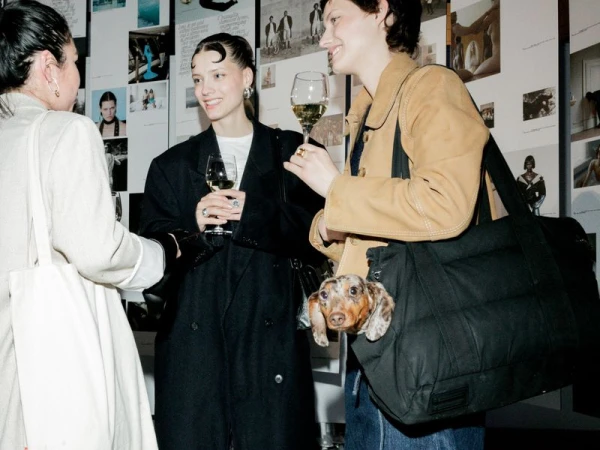 Three women converse at an event with a dachshund in a tote bag, illuminated by indoor lighting, creating a fashionable and social atmosphere.