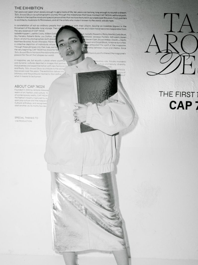 Adult female in studio standing in front of printed text, wearing a jacket and metallic skirt, holding a black book in her hands.