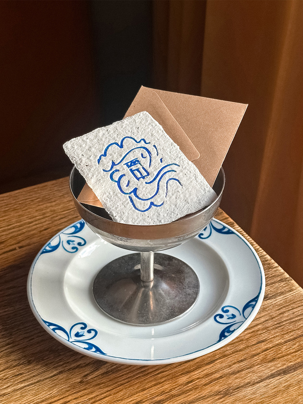 Textured paper card with blue ink illustration resting inside a metallic coupe glass on a blue and white patterned saucer.