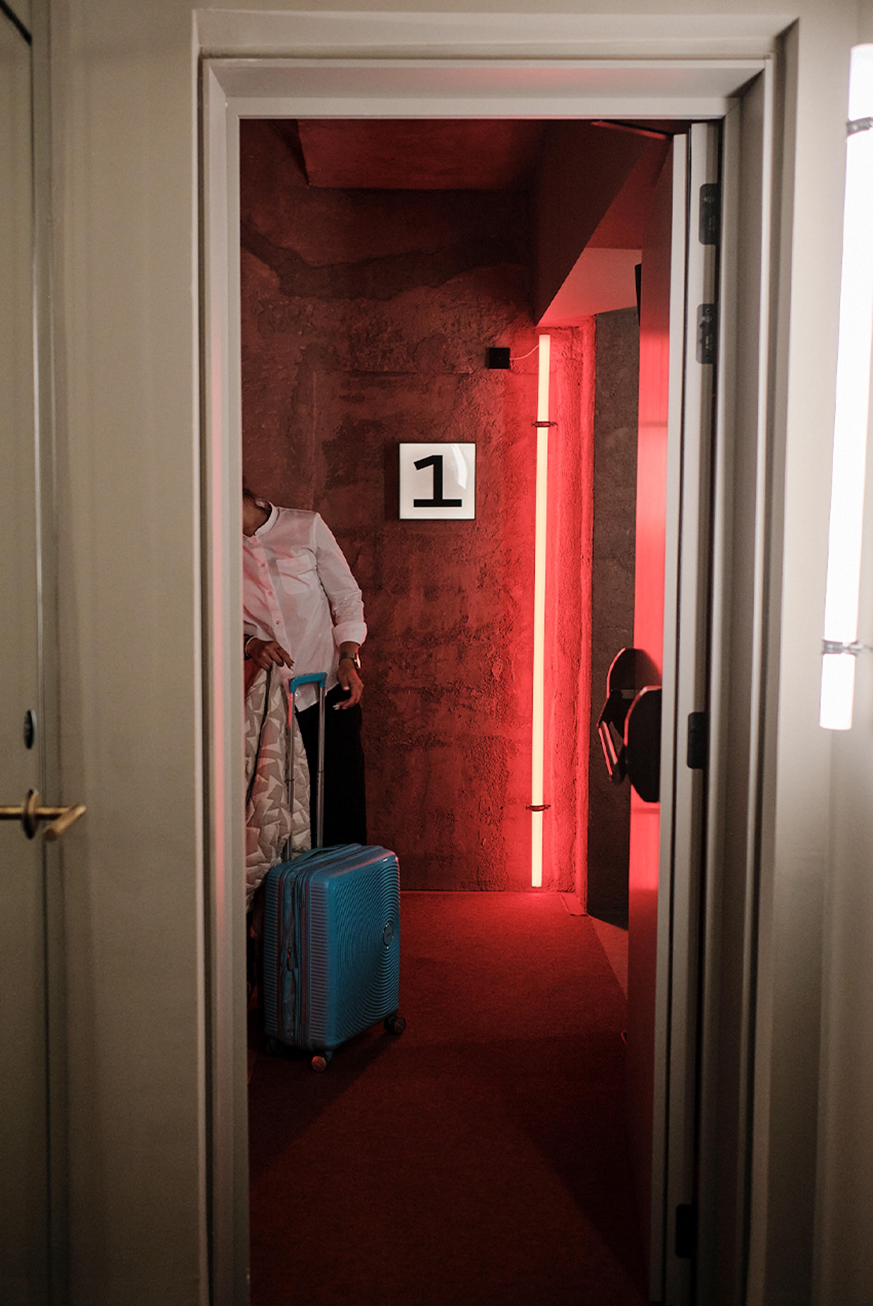 A person holding a patterned blanket standing next to a bright blue suitcase in a red lit hotel hallway