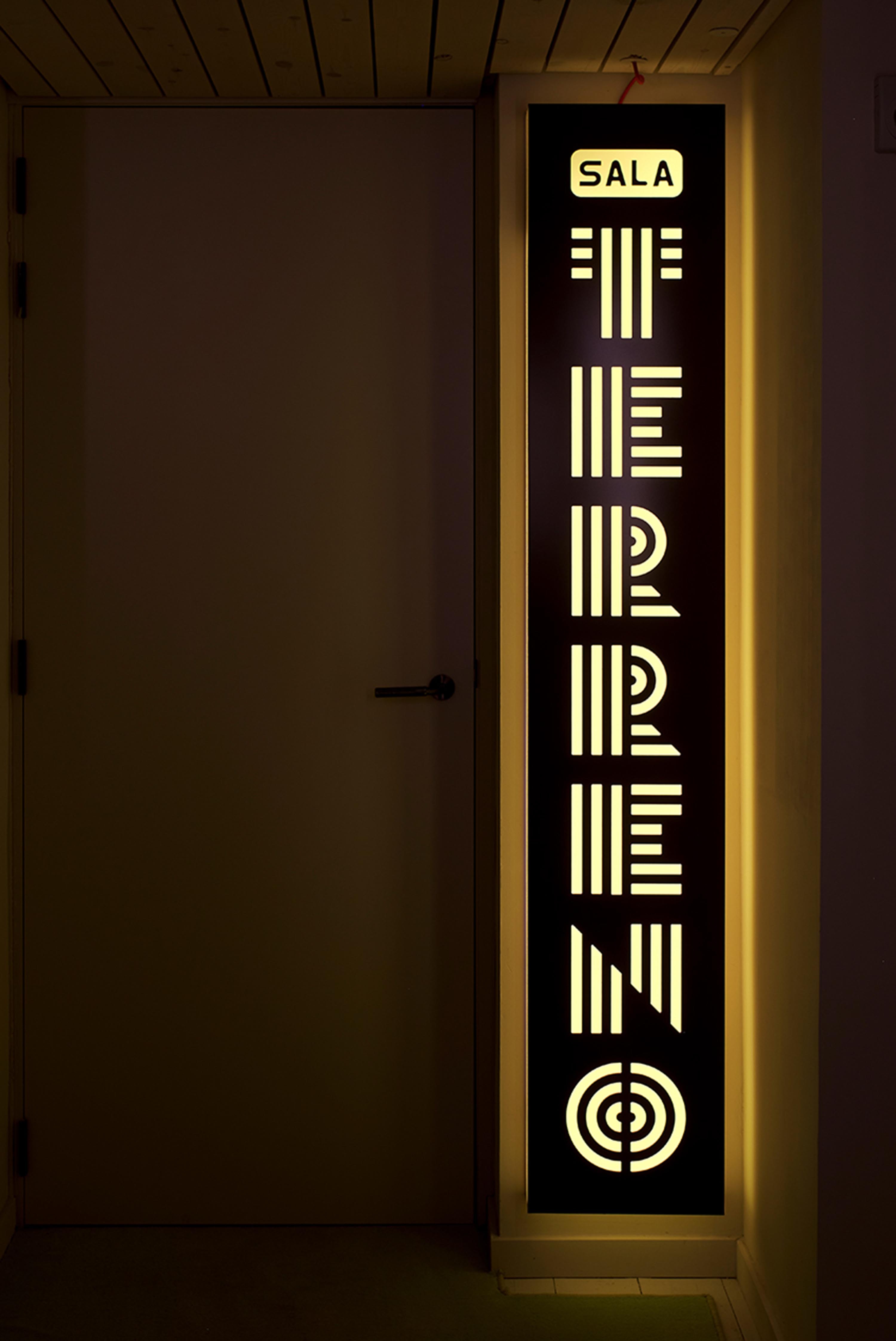 Vertical illuminated signage with geometric typography mounted on a wall next to a closed door.