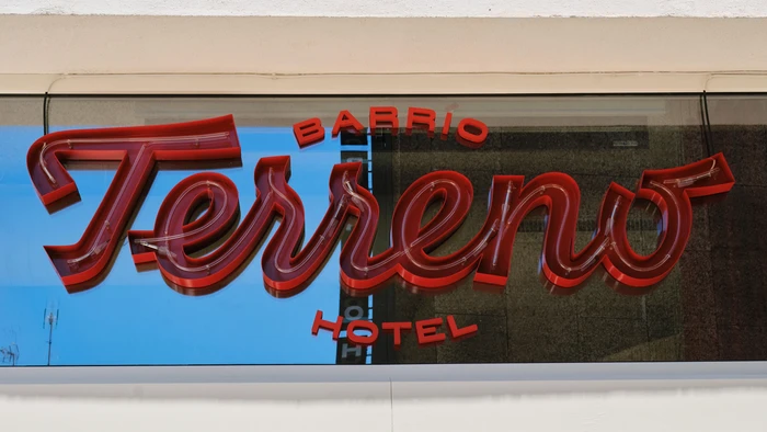 Red neon sign reading Barrio Terreno Hotel against a reflective glass facade and white architectural frame.