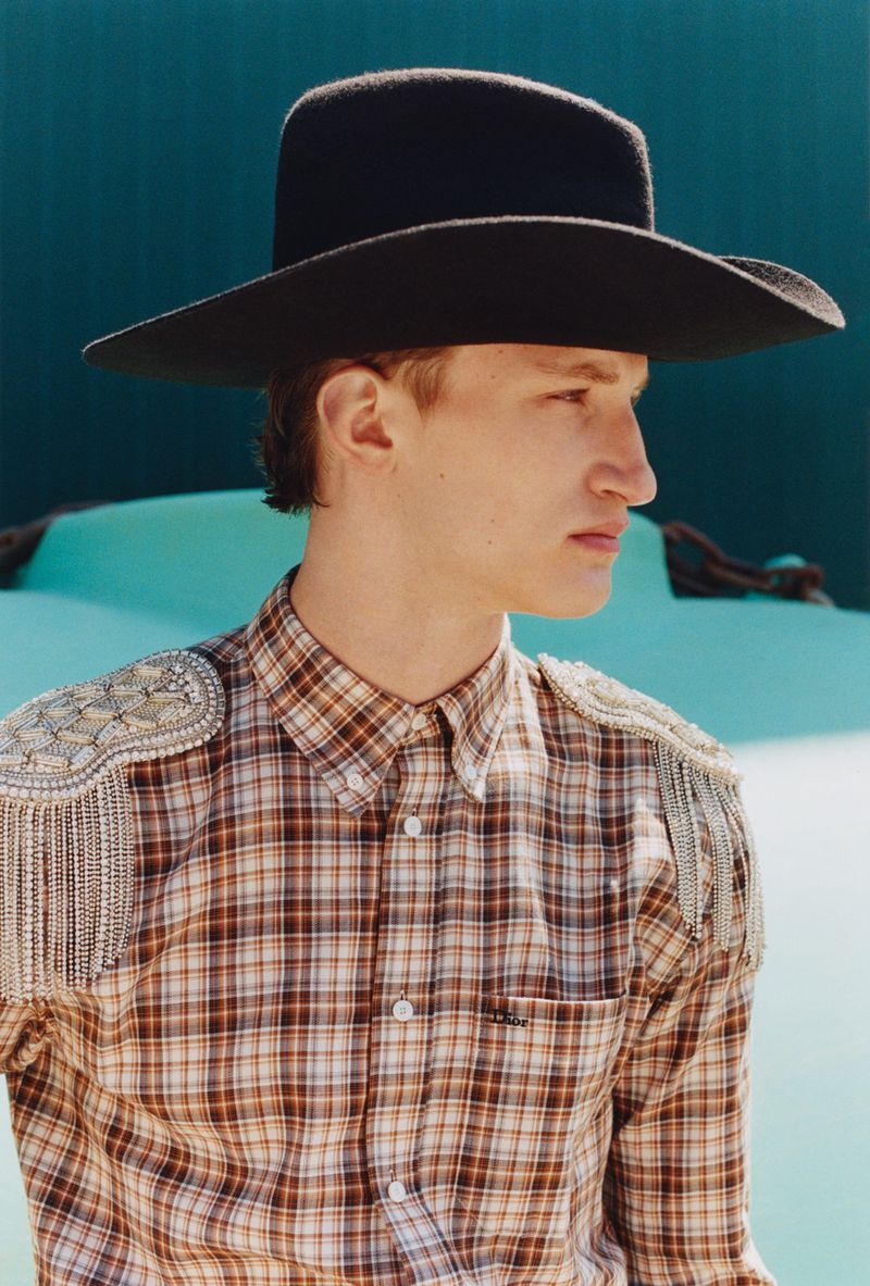 Model wearing a black cowboy hat and plaid shirt with embellished epaulets against a turquoise backdrop