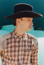 Model wearing a black cowboy hat and plaid shirt with embellished epaulets against a turquoise backdrop