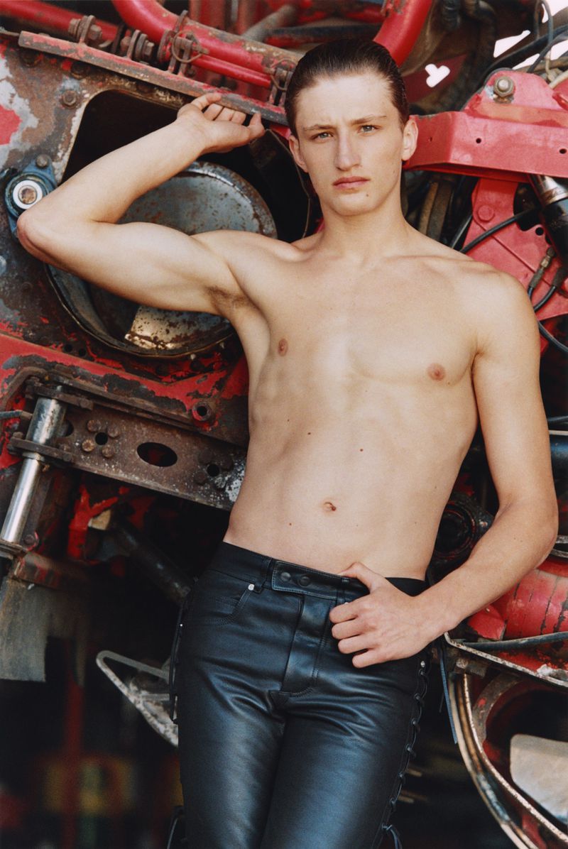 Shirtless male model wearing black leather trousers posing against heavy red industrial machinery