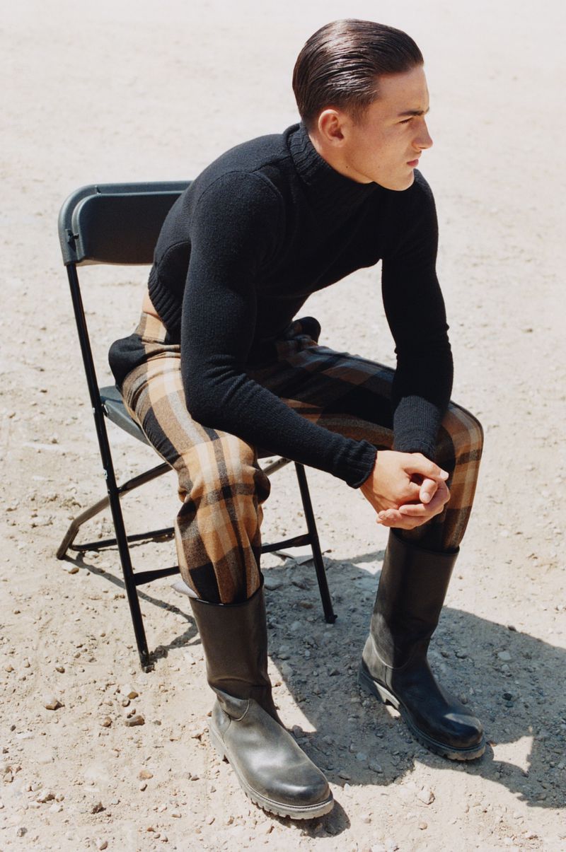 Model Lincoln Donald seated on a folding chair wearing a black turtleneck and plaid trousers in an outdoor desert setting.