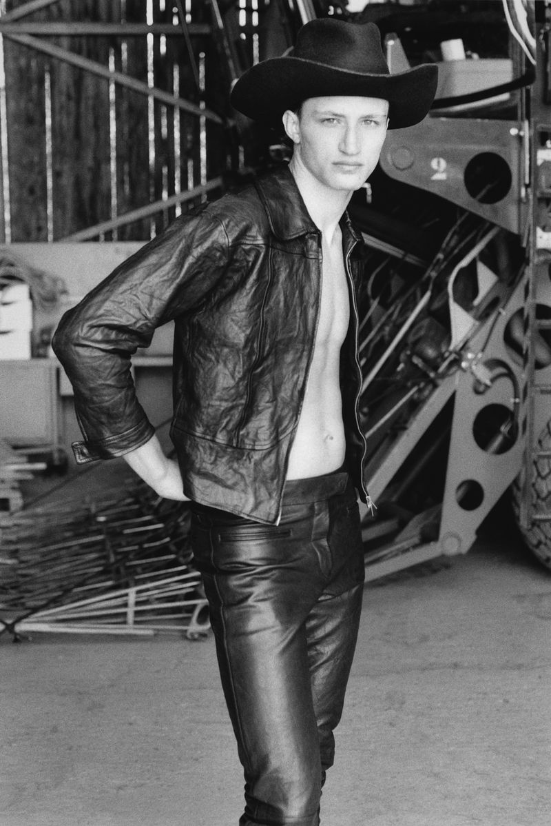 Black and white portrait of a male model wearing a black leather jacket, trousers, and cowboy hat outdoors.