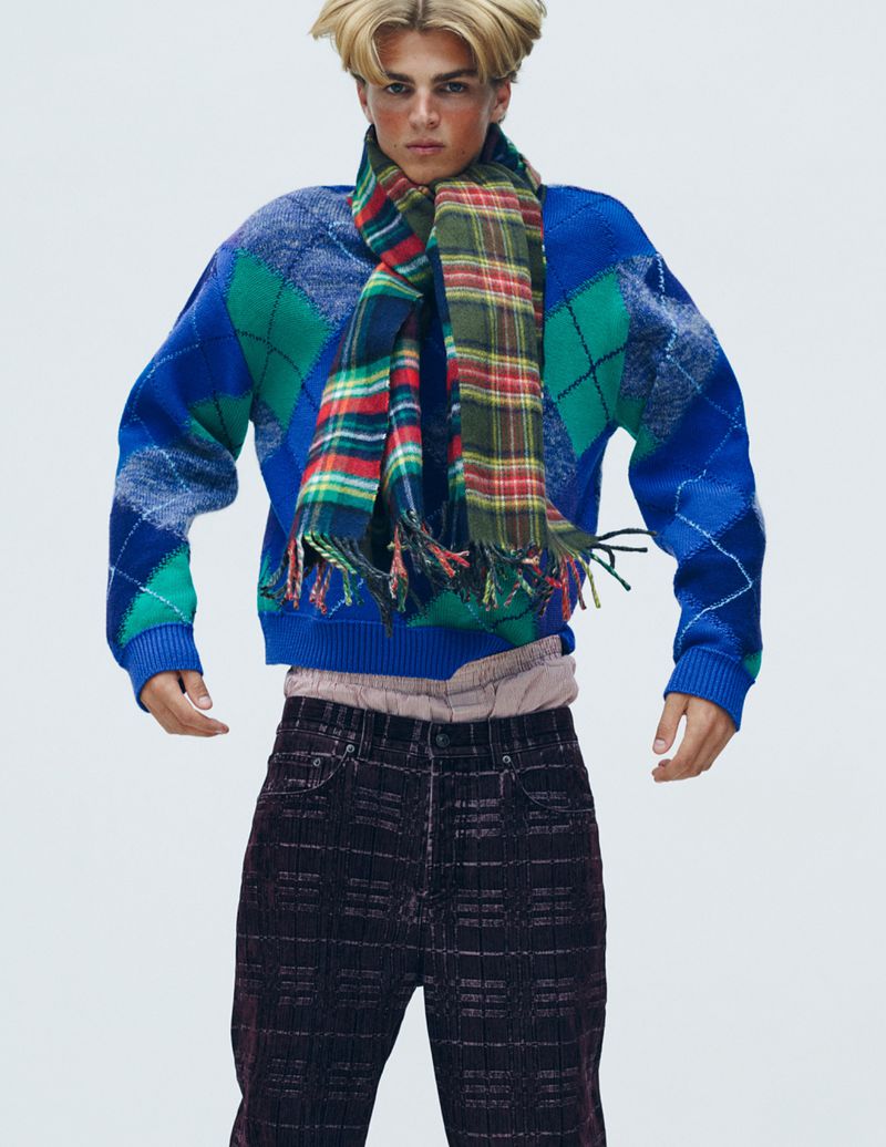 Male model wearing a blue argyle sweater and a plaid scarf against a seamless white background