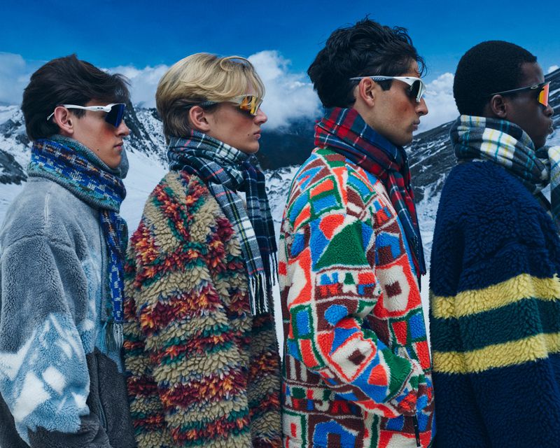 Four male models in patterned fleece jackets and scarves wearing athletic sunglasses outdoors in a snowy mountain landscape