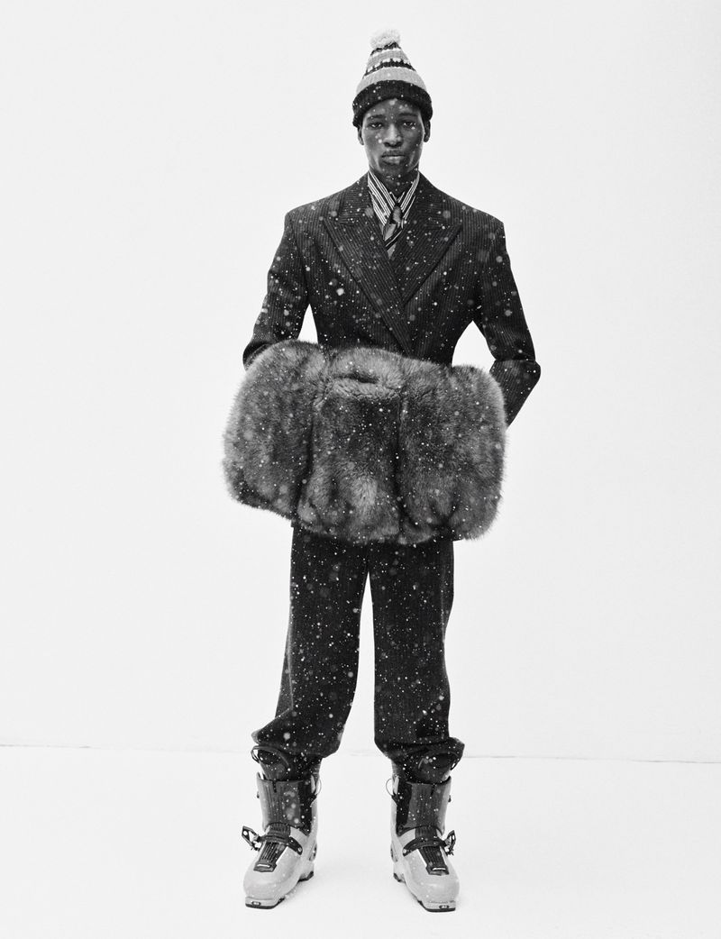 Model wearing a tailored suit, patterned beanie, and ski boots holding a large fur muff against a white backdrop.