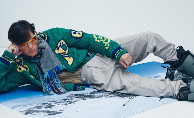 Model lying down wearing a green patterned sweater, grey trousers, and sports sunglasses against a white background.