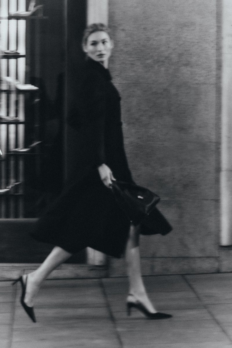 Grace Elizabeth walking in a black coat and heels holding a handbag past a storefront.
