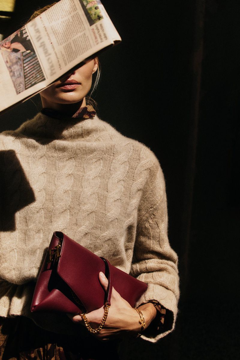 Model holding a burgundy leather clutch while partially obscuring her face with a newspaper under strong sunlight