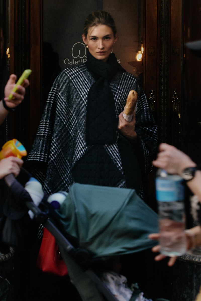Model Grace Elizabeth in a black and white plaid cape holding a baguette outside a cafe