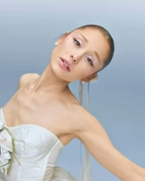 Ariana Grande wearing a white corset top with floral embellishments and pearl earrings against a light grey background