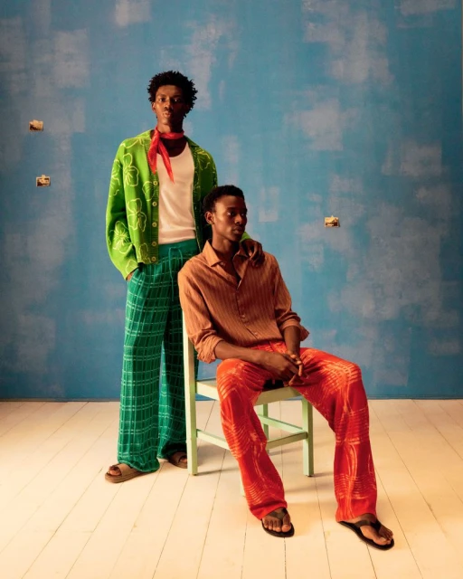Two African-descent adult men in casual summer fashion, one seated on a pale-green chair against a blue backdrop with painted textures.