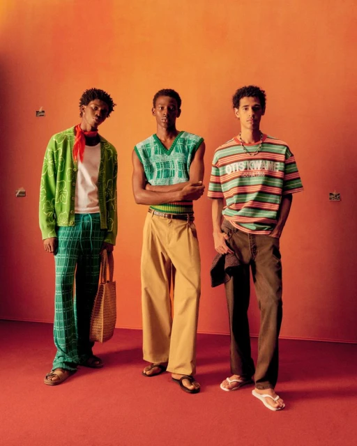 Three young Black men pose in a studio wearing stylish casual outfits, including a cardigan, vest, t-shirt, and pants against an orange backdrop.