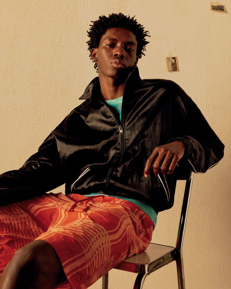Young Black male fashion model in black jacket and patterned orange shorts sitting on a metal chair against a neutral wall indoors.