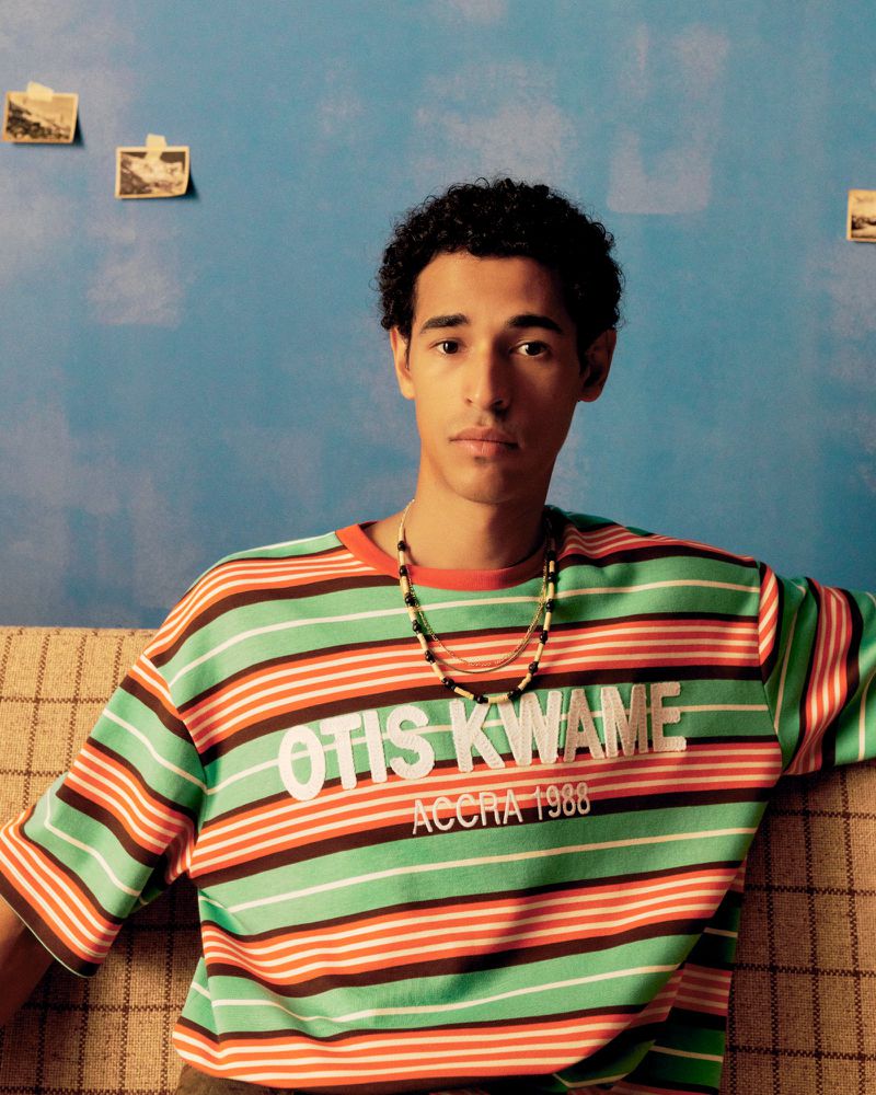 A young adult male is sitting indoors wearing a colorful striped shirt and beaded necklaces, posing against a blue textured wall.