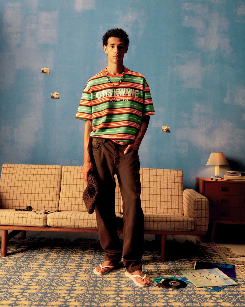 Young adult stands in a room with a striped shirt, holding a hat, with a couch, patterned rug, record albums, and a side table.