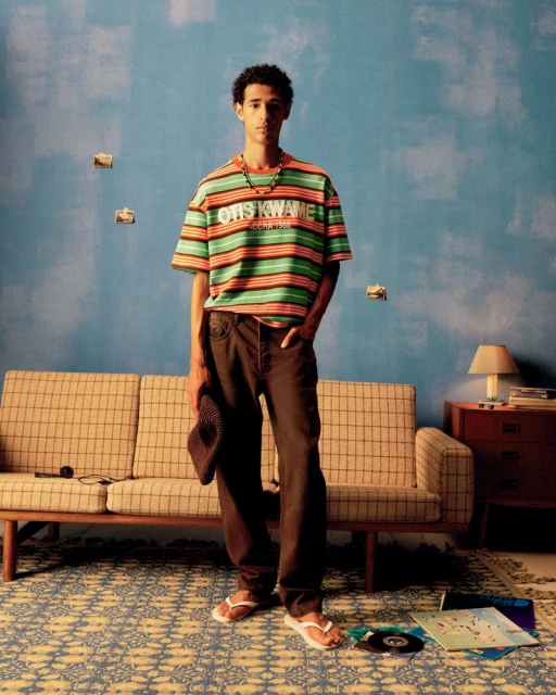 Young adult stands in a room with a striped shirt, holding a hat, with a couch, patterned rug, record albums, and a side table.