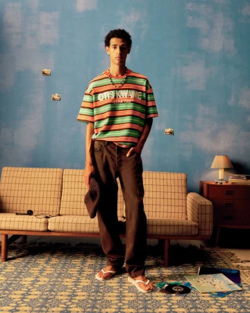 Young adult stands in a room with a striped shirt, holding a hat, with a couch, patterned rug, record albums, and a side table.