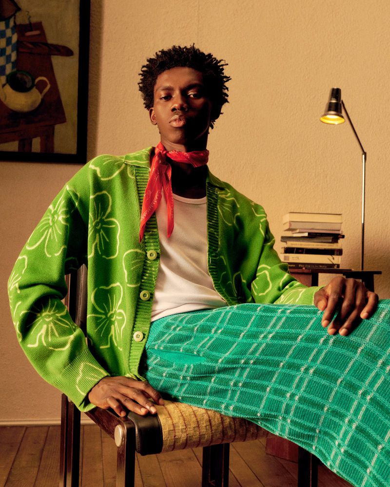 Adult male poses in a green floral cardigan and plaid pants while sitting on a chair in a brightly lit indoor setting.
