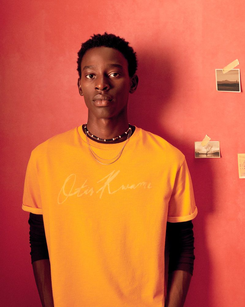 Portrait of a young adult male in yellow t-shirt against a warm-toned wall with small pictures. Fashion, beauty, and lifestyle.