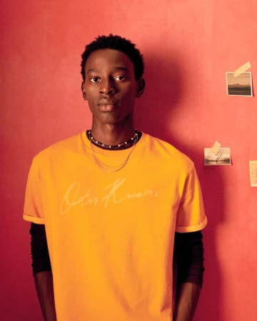 Portrait of a young adult male in yellow t-shirt against a warm-toned wall with small pictures. Fashion, beauty, and lifestyle.