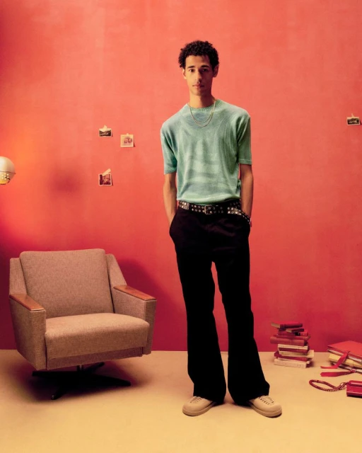 Full shot of a young adult man in a light blue shirt and black pants, posing in a photo studio with retro decor, looking at the camera.