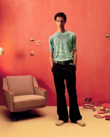 Full shot of a young adult man in a light blue shirt and black pants, posing in a photo studio with retro decor, looking at the camera.