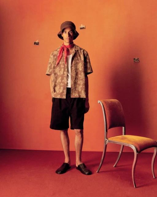 Adult male poses in a patterned shirt, dark shorts, bucket hat, and scarf in a studio. A chair sits to the right of him.