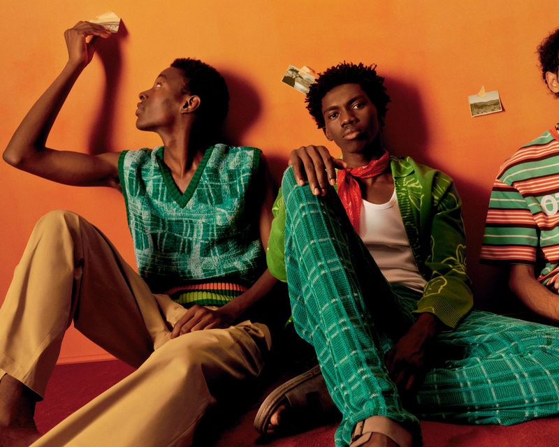 Three African-descent men seated on an orange backdrop in a photo studio wearing 70's-inspired patterned casual wear.