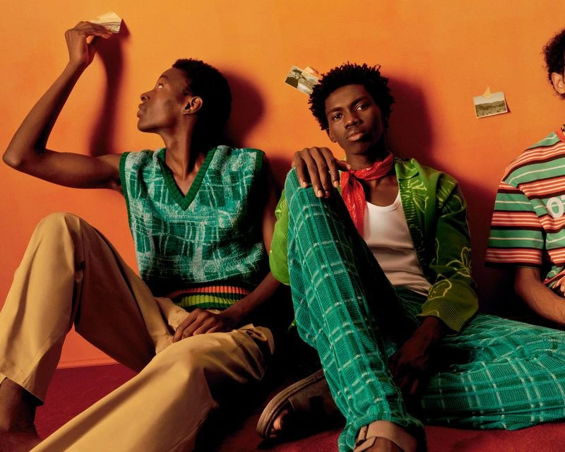 Three African-descent men seated on an orange backdrop in a photo studio wearing 70's-inspired patterned casual wear.