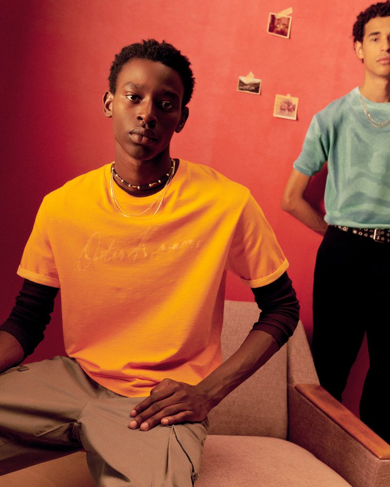 Two men pose indoors against a red wall with attached photos, one seated in a neutral chair wearing yellow t-shirt and cargo pants.