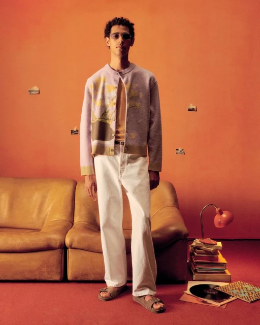 A slender adult male wearing a light patterned cardigan, white jeans, and sandals poses in front of an orange backdrop with a sofa.