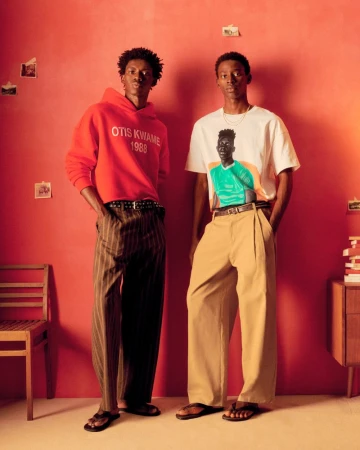 Two Black men in stylish streetwear fashion stand against a red wall indoors, showcasing casual attire and a retro, fashion-forward aesthetic.