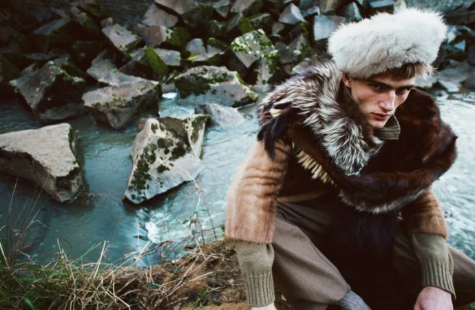 Male model wearing a fur hat and fur wrap coat crouching by a rocky riverbank in HERO Magazine editorial.