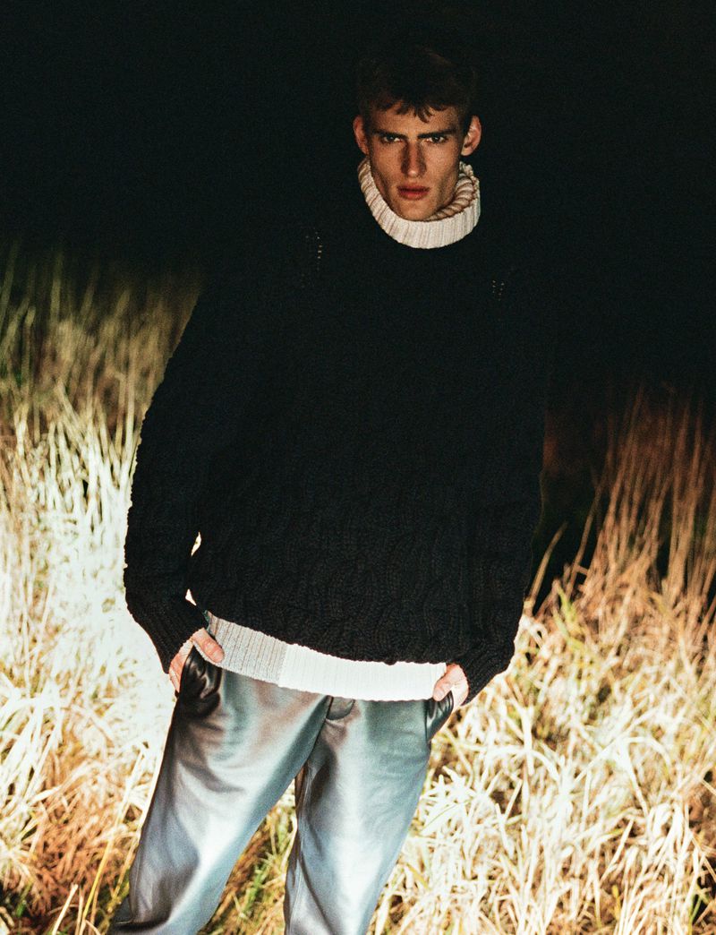 Young male model in dark sweater and metallic trousers standing in a grassy field at night with flash lighting.