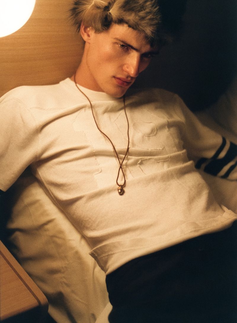 A young man wearing a furry hat and a white t-shirt with a pendant necklace relaxing indoors.