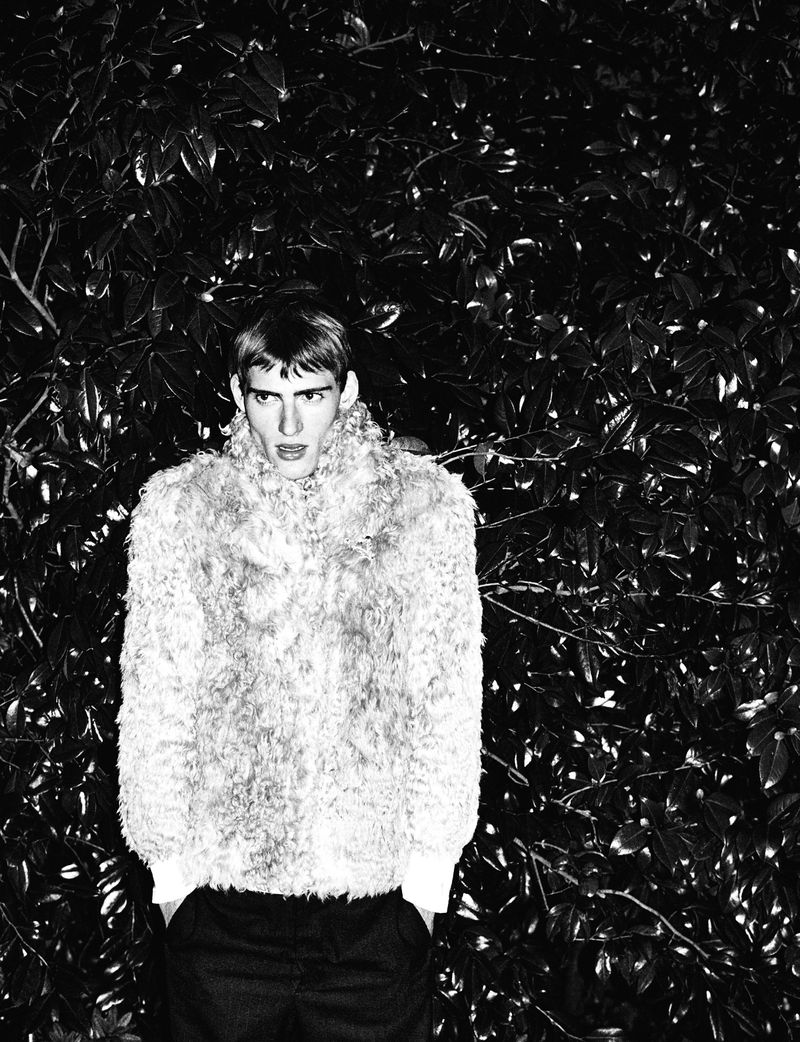 Black and white portrait of a male model wearing a textured faux fur jacket against dark foliage for HERO Magazine.