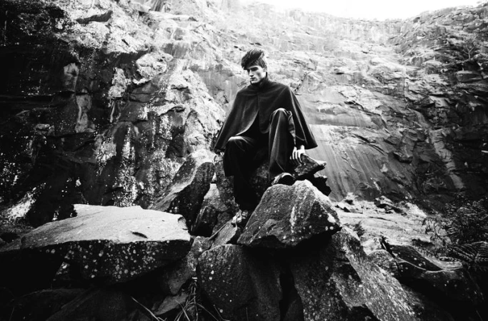 A monochrome fashion editorial image of a male model perched on a rocky outcrop amidst a rugged stone quarry.