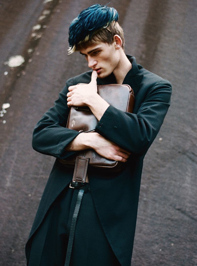 Young man with dark blue hair holding a brown leather briefcase against an outdoor asphalt background