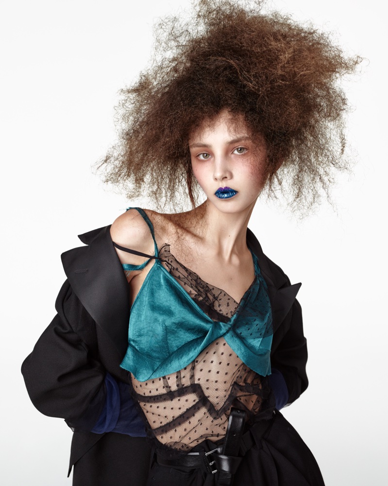 Model with voluminous textured hair wearing a teal satin camisole and black jacket against a white background