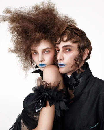 Portrait of two models with elaborate hairstyles and blue lipstick wearing black garments against a white background