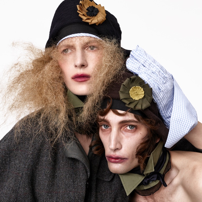 Two models wearing dark hats adorned with fabric rosette flowers and layered textured garments against a plain white background