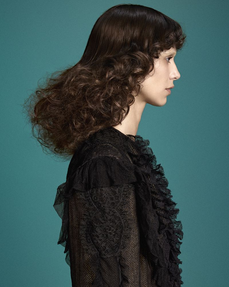 Profile portrait of a woman with voluminous dark brown curly hair wearing a black lace ruffled dress against a teal background.