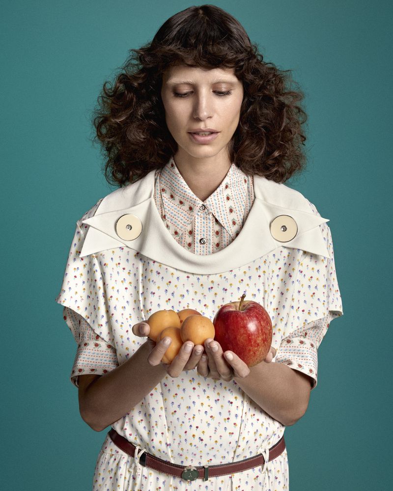 Model with dark curly hair holding small apricots and a red apple against a teal background
