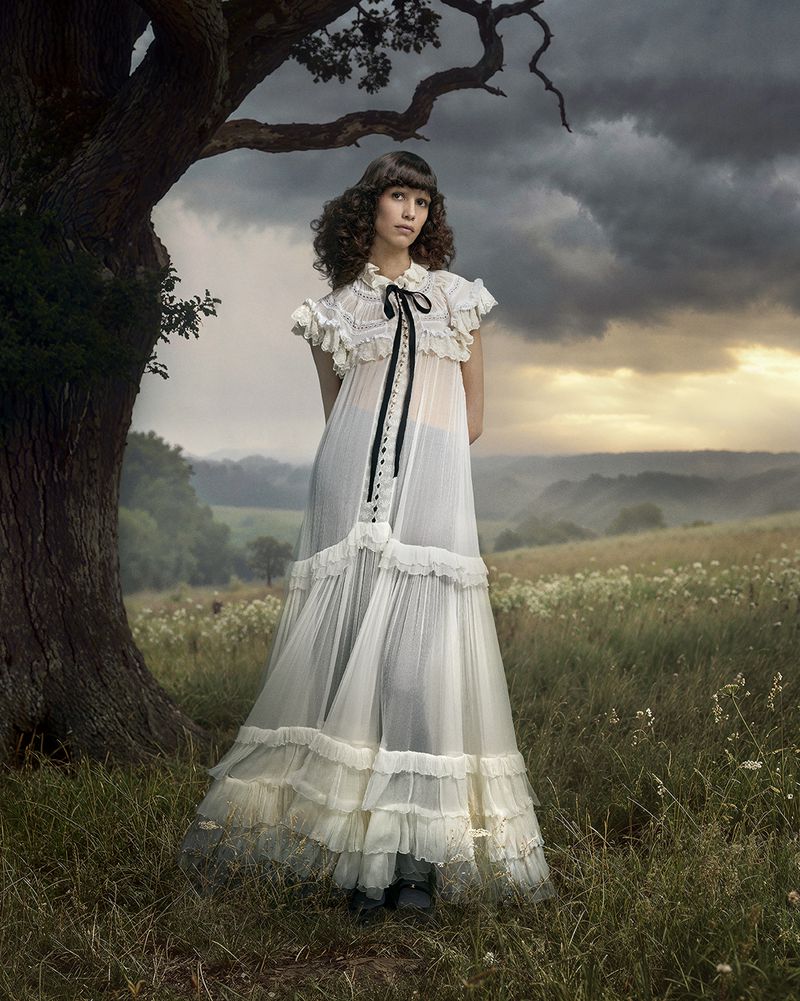 Model standing beneath a large gnarled tree in an open meadow wearing a sheer white ruffled gown with black ribbon detailing.