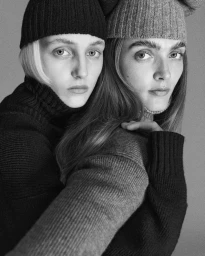 Two female models wearing ribbed knit beanies and turtleneck sweaters posed together in a studio portrait.